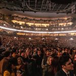 Grande público reunido em uma arena durante um evento musical, sob luzes intensas.