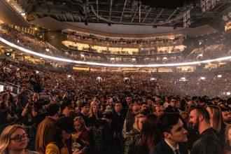 Grande público reunido em uma arena durante um evento musical, sob luzes intensas.