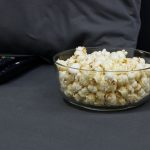 Tigela de vidro com pipoca ao lado de controles remotos de TV sobre um sofá cinza, em clima de noite de cinema em casa.