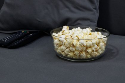 Tigela de vidro com pipoca ao lado de controles remotos de TV sobre um sofá cinza, em clima de noite de cinema em casa.