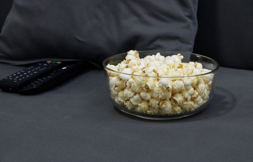 Tigela de vidro com pipoca ao lado de controles remotos de TV sobre um sofá cinza, em clima de noite de cinema em casa.