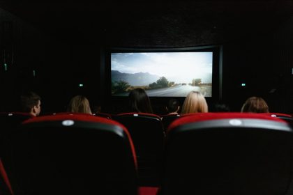 Espectadores vistos de costas assistem a um filme em uma sala de cinema com poltronas vermelhas.