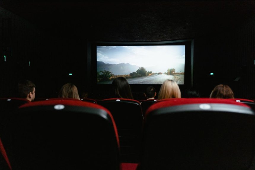 Espectadores vistos de costas assistem a um filme em uma sala de cinema com poltronas vermelhas.