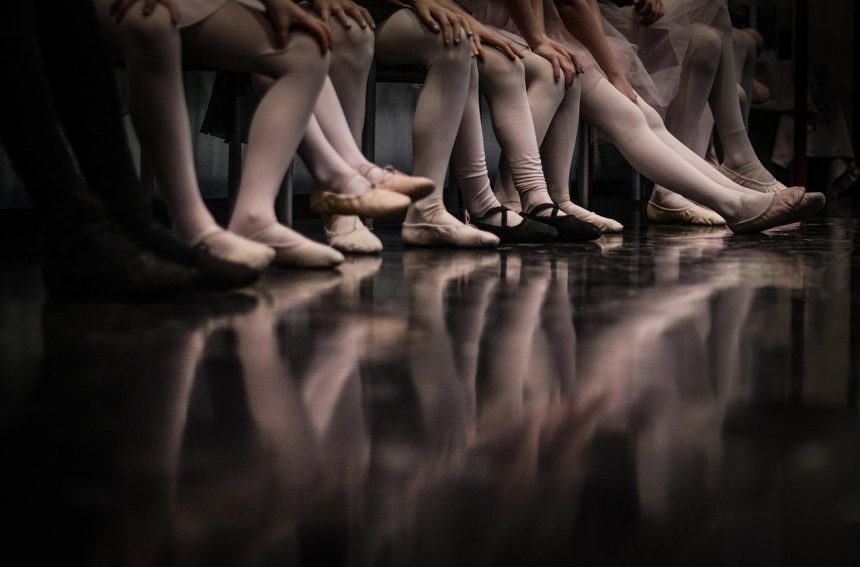 Bailarinas se apresentam em um palco de balé, com os pés refletidos no piso.