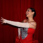 Mulher com vestido vermelho brilhante se apresenta em um palco diante de cortinas vermelhas.