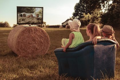 Família reunida ao ar livre, sentada sobre fardos de palha em um campo, assistindo televisão.