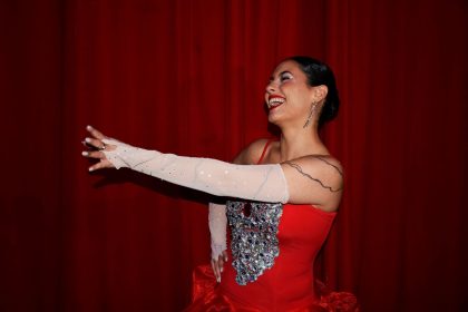 Mulher com vestido vermelho brilhante se apresenta em um palco diante de cortinas vermelhas.