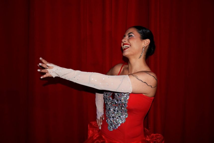 Mulher com vestido vermelho brilhante se apresenta em um palco diante de cortinas vermelhas.